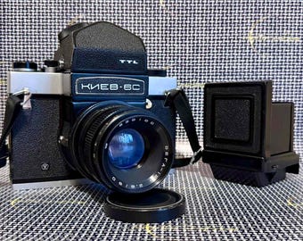 May include: A black and silver Kiev-6C TTL medium format film camera with a Vega-12B 2.8/90 lens attached. The camera features a prominent prism viewfinder, a black strap, and a separate detachable film back accessory placed to the right.