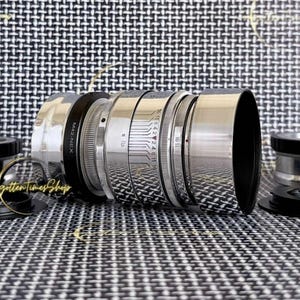 May include: A silver camera lens with a black lens hood, featuring engraved aperture markings and focus rings. Two metal lens adapters are positioned on either side, one labelled M42-M4/3 and the other labelled M42-FX, with an M42-NEX adapter attached.