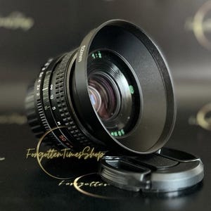 May include: A black camera lens with a 52mm filter thread and a black lens hood. The lens has a depth of field scale and aperture markings.