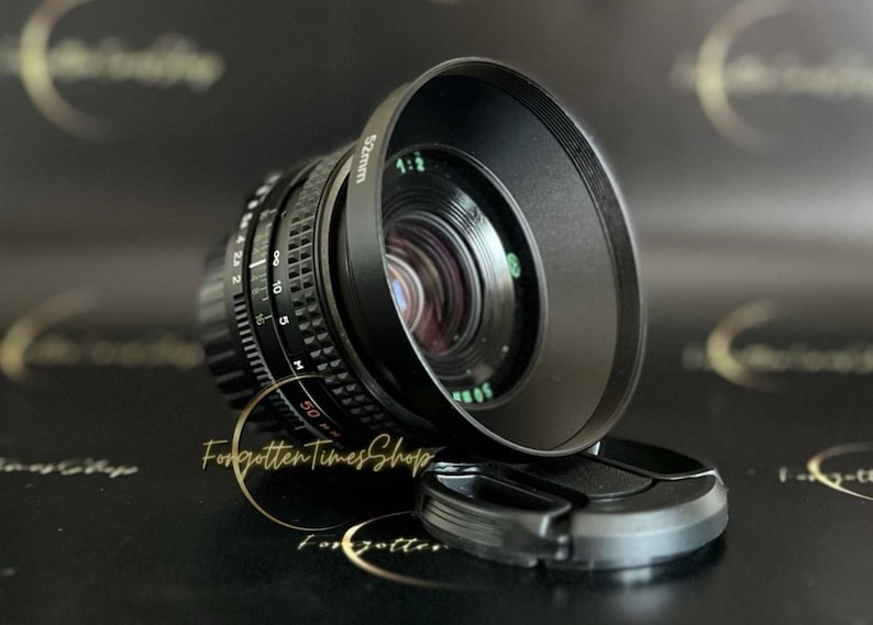 May include: A black camera lens with a 52mm filter thread and a black lens hood. The lens has a depth of field scale and aperture markings.