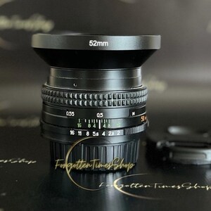 May include: A black camera lens with a 52mm filter thread. The lens has a black metal body and a silver ring with markings for aperture and distance. The lens is sitting on a black surface.