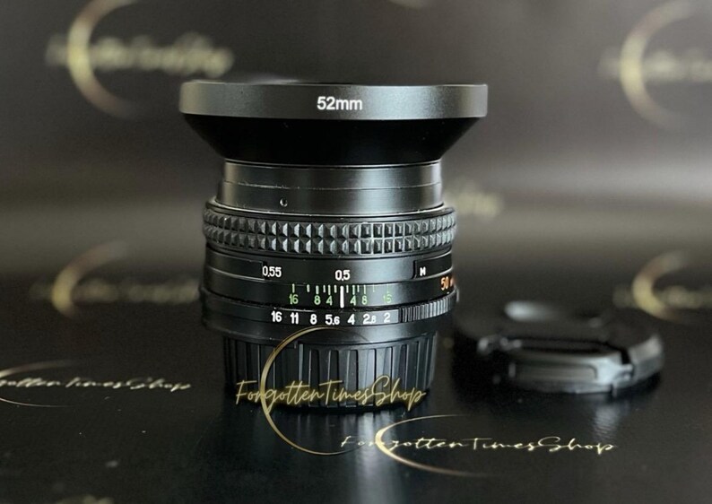 May include: A black camera lens with a 52mm filter thread. The lens has a black metal body and a silver ring with markings for aperture and distance. The lens is sitting on a black surface.