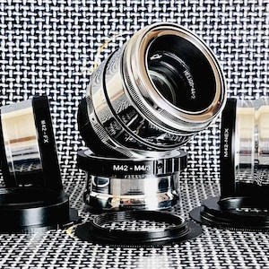 May include: A collection of camera lens adapters and a lens. The lens is silver and black with the text "HELIOS-44-2". The adapters are black and have text such as "M42-FX", "M42-M4/3", and "M42-NEX".