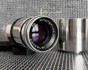May include: A silver-toned camera lens with the text "ЮПИТЕР-IIA 4/85" and a matching silver cylindrical container. The lens has a black ring with white text and a black lens cap. The background is a black and white checkered pattern.