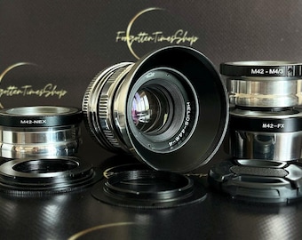 May include: A silver camera lens with a black lens cap and a black and silver adapter ring. The lens has the text "HELIOS-44M-4" printed on it. The adapter rings have the text "M42-NEX", "M42-M4/3", and "M42-FX" printed on them.