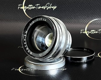 May include: A vintage silver and black camera lens with the text "ЮПИТЕР-8 M" and "F=5 CM" visible. The lens has a black cap and is resting on a silver base. The background is black with gold text.