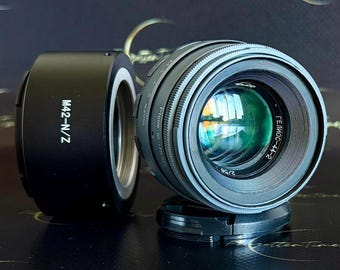 May include: A black M42-N/Z lens adapter next to a silver and black camera lens. The lens has the text "Гелиос-44-2" and "2/58" printed on it. The lens is resting on a black surface.