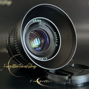 May include: A black camera lens with a silver ring around the edge. The lens has the text "ARSATH" and "50 mm 2.1" printed on it.
