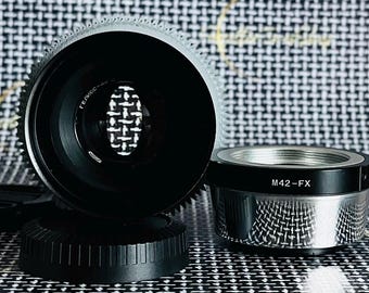 May include: A black camera lens with a silver adapter ring. The lens has a black metal body and a silver metal ring with the text "M42-FX" printed on it.