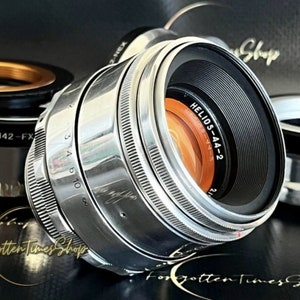May include: A silver camera lens with the text "HELIOS-44-2" printed on the side. The lens has a black aperture ring with markings for f-stops 2.0, 2.8, 4, 5.6, 8, 11, and 16. The lens is in focus and the background is blurred.