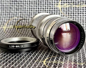 May include: A silver Jupiter-11 4/135 camera lens with a purple-tinted front glass element and a black L39-M4/3 adapter ring. The lens barrel features engraved aperture markings and knurled metal focus rings, showcasing a metallic finish.