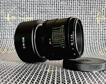 May include: Black camera lens with a lens hood and a lens cap. The lens has markings for aperture and focus, with the text "M42-N/Z" visible. The lens cap is black and round.