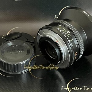 May include: A black Nikon camera lens with a 50mm focal length and a silver aperture ring. The lens has a black lens cap with the Nikon logo.