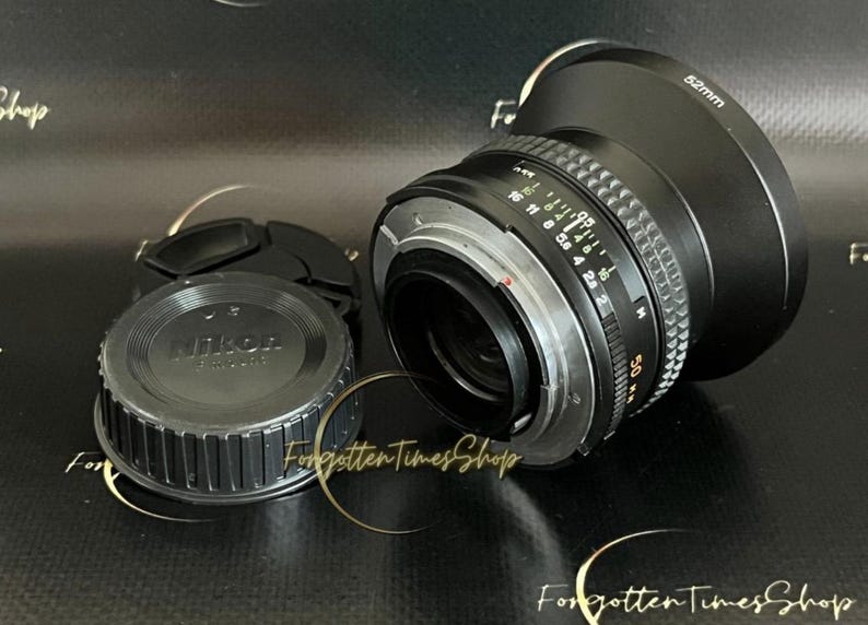 May include: A black Nikon camera lens with a 50mm focal length and a silver aperture ring. The lens has a black lens cap with the Nikon logo.