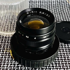 May include: A black MIR-1 2.8/37 camera lens with a clear plastic case and a black lens cap. The lens has a gold-colored interior and white text. The lens is sitting on a black base.