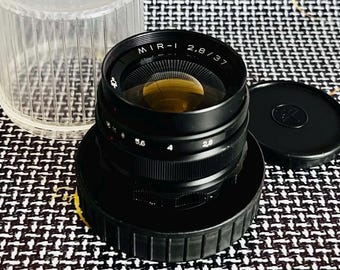 May include: A black MIR-1 2.8/37 camera lens with a clear plastic case and a black lens cap. The lens has a gold-colored interior and white text. The lens is sitting on a black base.