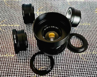 May include: A collection of black camera lenses and adapters, including a MIR-1 2.8/37 lens, various M42-NEX and M42-M4/3 adapters, and lens rings. The items are arranged on a patterned surface.