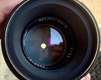 May include: A black Helios-44-2 2/58 camera lens featuring a circular glass element with visible aperture blades inside. The front rim displays the text HELIOS-44-2, 2/58, and the serial number 7205187 in white lettering against the black metal housing.
