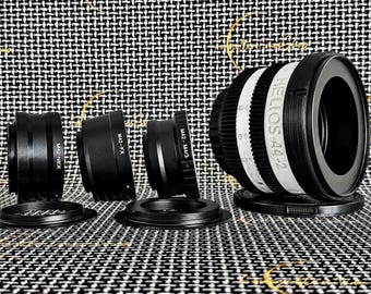 May include: A black Helios-44-2 camera lens with a white focus ring featuring black text and markings, accompanied by three black metal lens adapters labeled M42-NEX, M42-FX, and M42-M4/3, each positioned with its respective black circular lens cap.