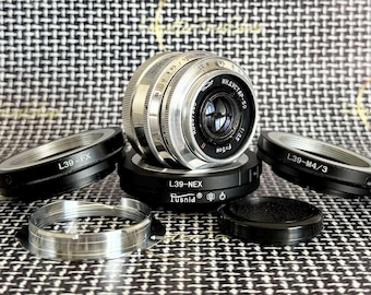 May include: A silver Industar-50 3.5/50 camera lens with Cyrillic text, mounted on a black L39-NEX adapter. Included are two additional black lens adapters labeled L39-FX and L39-M4/3, a separate silver metal ring, and a small black lens cap.