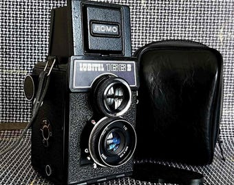 May include: A black Lubitel 166B twin-lens reflex camera with a leather case. The camera has two lenses, a top viewfinder, and the word "LOMO" on top. The camera is on a patterned surface.