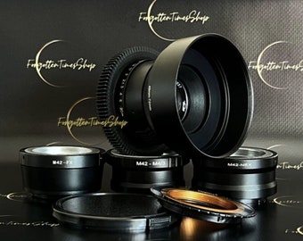 May include: A set of five black camera lens adapters with different mount types, including M42-FX, M42-M4/3, and M42-NEX. The adapters are arranged on a black surface with a lens hood in the background.