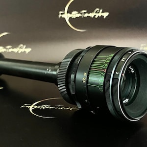 Puede incluir: Un objetivo negro y verde con un anillo plateado y marcas. El objetivo tiene un cuerpo de metal negro y un anillo plateado alrededor del frente. El objetivo está marcado con los números 2, 2.8, 4, 5.6, 8, 11, 16, 22 y 32. El objetivo también está marcado con las letras "M 0.5".