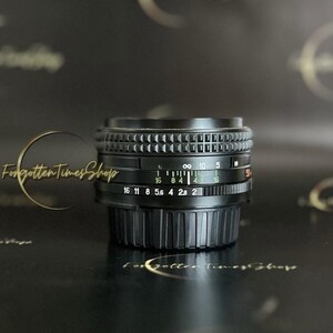 May include: A black camera lens with a silver ring and a depth of field scale. The lens has markings for f-stops from 16 to 2 and a distance scale from 0.5 metres to infinity.