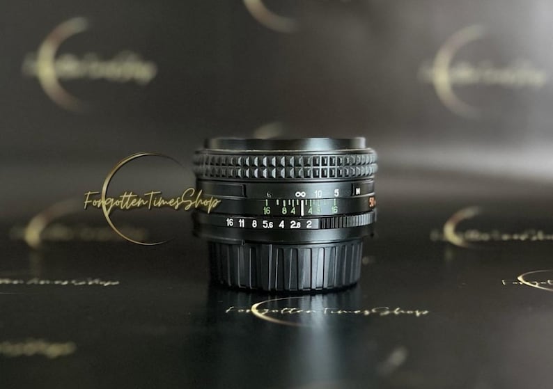 May include: A black camera lens with a silver ring and a depth of field scale. The lens has markings for f-stops from 16 to 2 and a distance scale from 0.5 metres to infinity.