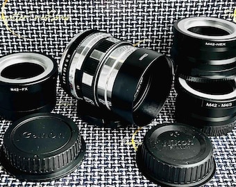 May include: A collection of camera lens adapters, including a Canon lens adapter, a Nikon lens adapter, and a M42-NEX lens adapter. The adapters are black and silver and have various markings, including "M42-FX", "M42 - M4/3", and "M42-NEX".