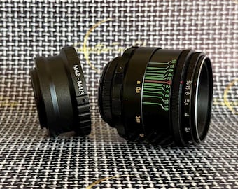 May include: A black camera lens and a separate M42 to M4/3 lens adapter. The lens features a black barrel with white and green numerical markings for aperture and distance settings, including values like 2.8, 4, 5.6, 8, 11, and 16.