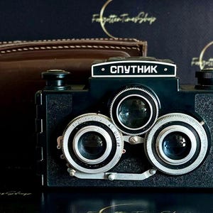 May include: A black vintage Sputnik stereo camera with three lenses and the word "Спутник" in white lettering. A brown leather case sits behind the camera. The camera has silver dials and a retro design.