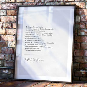 May include: Framed print featuring a quote by Ralph Waldo Emerson. The text is in black on a white background, set within a black frame. The quote is about success and is displayed against a brick wall and wooden floor.