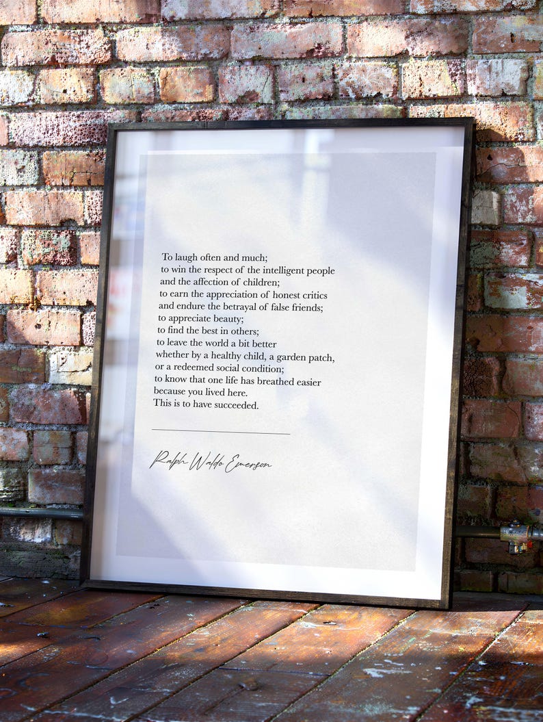May include: Framed print featuring a quote by Ralph Waldo Emerson. The text is in black on a white background, set within a black frame. The quote is about success and is displayed against a brick wall and wooden floor.