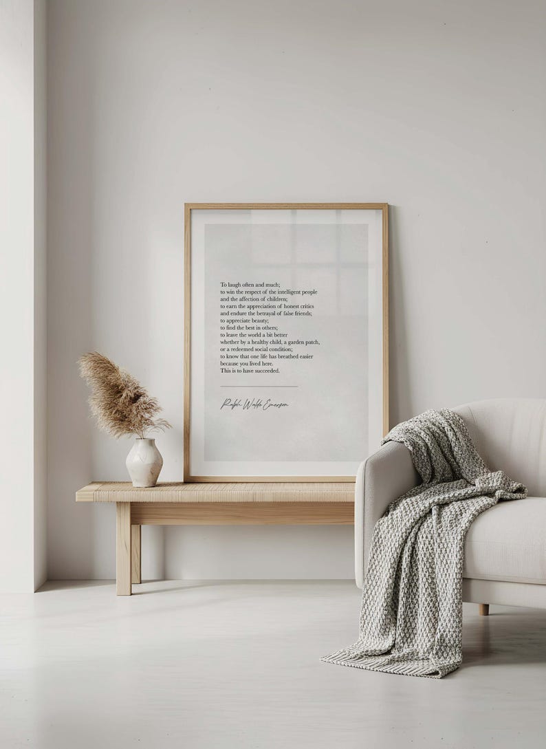 May include: A framed print with a light wood frame displays a poem in black text. The print rests on a light wood bench with a white vase and dried flowers. A white sofa with a gray blanket is to the right.
