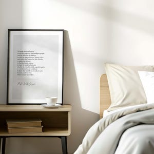 May include: A framed print with a black border displays a poem in a minimalist style. The print rests on a wooden nightstand with a cup and saucer, and two books. A bed with neutral-toned bedding is in the background.