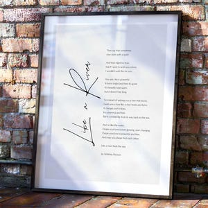 May include: Framed print with a poem about love, featuring the words "Like a River" in cursive. The poem is in black text on a white background, set within a black frame. The background is a brick wall.
