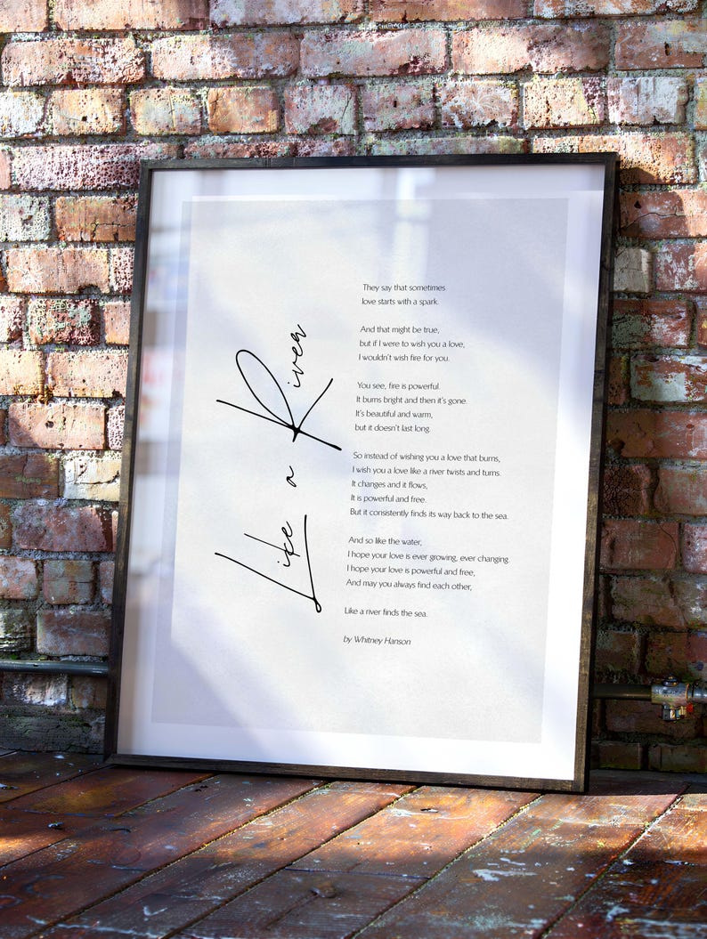 May include: Framed print with a poem about love, featuring the words "Like a River" in cursive. The poem is in black text on a white background, set within a black frame. The background is a brick wall.