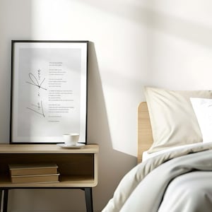 May include: A framed black and white print with a poem and abstract design sits on a wooden nightstand. A white teacup and saucer are on the nightstand, with two books below. A bed with a beige headboard and pillows is in the background.