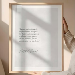 May include: A framed print with a light wood frame. The print features a quote by Viktor E. Frankl in black text on a white background. The quote reads, "Between stimulus and response there is a space..." The print is held by a person.