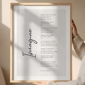 Puede incluir: Una impresión enmarcada con la letra de la canción "Imagine" de John Lennon. El texto está en una fuente sencilla sobre un fondo claro. La palabra "Imagine" está escrita en cursiva. El marco es de color madera clara.