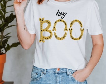 May include: White t-shirt with the text "hey BOO" in gold lettering with black accents. The letters are decorated with small black leaves and dots.