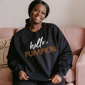 May include: A black sweatshirt with the text "hello, PUMPKIN" in white and orange lettering. The sweatshirt is being worn by a person sitting on a pink couch.