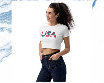May include: A white cropped t-shirt with the word "USA" printed on the front. The letters are made up of red, white, and blue stripes and stars, resembling the American flag.