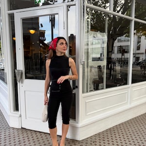 Può includere: Un top nero senza maniche a collo alto abbinato a pantaloni capri neri, una bandana rossa a fantasia tra i capelli e sandali neri a punta aperta. Una piccola borsa beige è tenuta in mano e un braccialetto dorato è indossato al polso.
