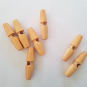 Peut inclure: Plusieurs boutons en bois de couleur brun clair. Chaque bouton a une forme cylindrique avec un haut et un bas effilés, et une fente horizontale au milieu. Les boutons sont dispersés sur une surface blanche.