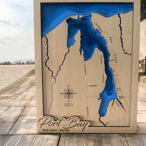 May include: A wooden framed 3D map of Port Bay, Wayne County, New York. The map features layered blue water depths and etched roads. The text "Port Bay" and coordinates are visible.