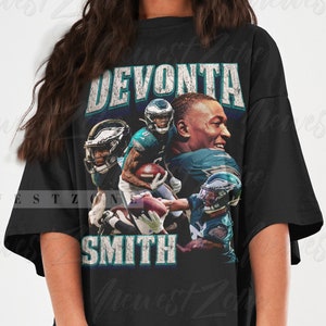 May include: Black t-shirt featuring a graphic of football player DeVonta Smith. The design includes the name "DEVONTA SMITH" in a distressed, vintage style. The shirt showcases images of the player in action, with the Philadelphia Eagles logo.