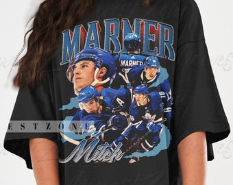 May include: Black t-shirt featuring a collage of hockey player images in blue and white, with the name "MARNER" in a vintage font. The player's name "Mitch" is also displayed in a script font. The shirt is a casual, oversized style.