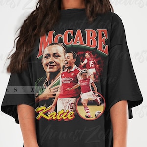 Könnte beinhalten: Schwarzes Oversize-T-Shirt mit einem Vintage-Grafikdruck einer weiblichen Fußballspielerin in einem rot-weißen Trikot. Die Grafik enthält den Text "MCCABE" und "Katie".